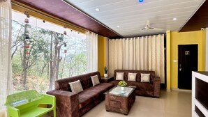 Una sala de estar con dos sofás, una mesita de centro y una silla verde. Hay una ventana con cortinas y vistas a árboles.