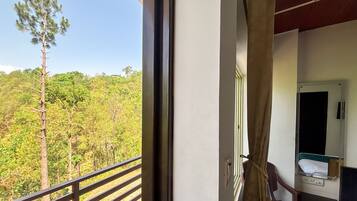 A room with a window overlooking a forested area, a bed, and a small table with a chair.