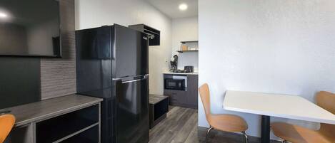 Cocina compacta con nevera negra, microondas y cafetera. Cuenta con un pequeño comedor con una mesa blanca y dos sillas de madera.