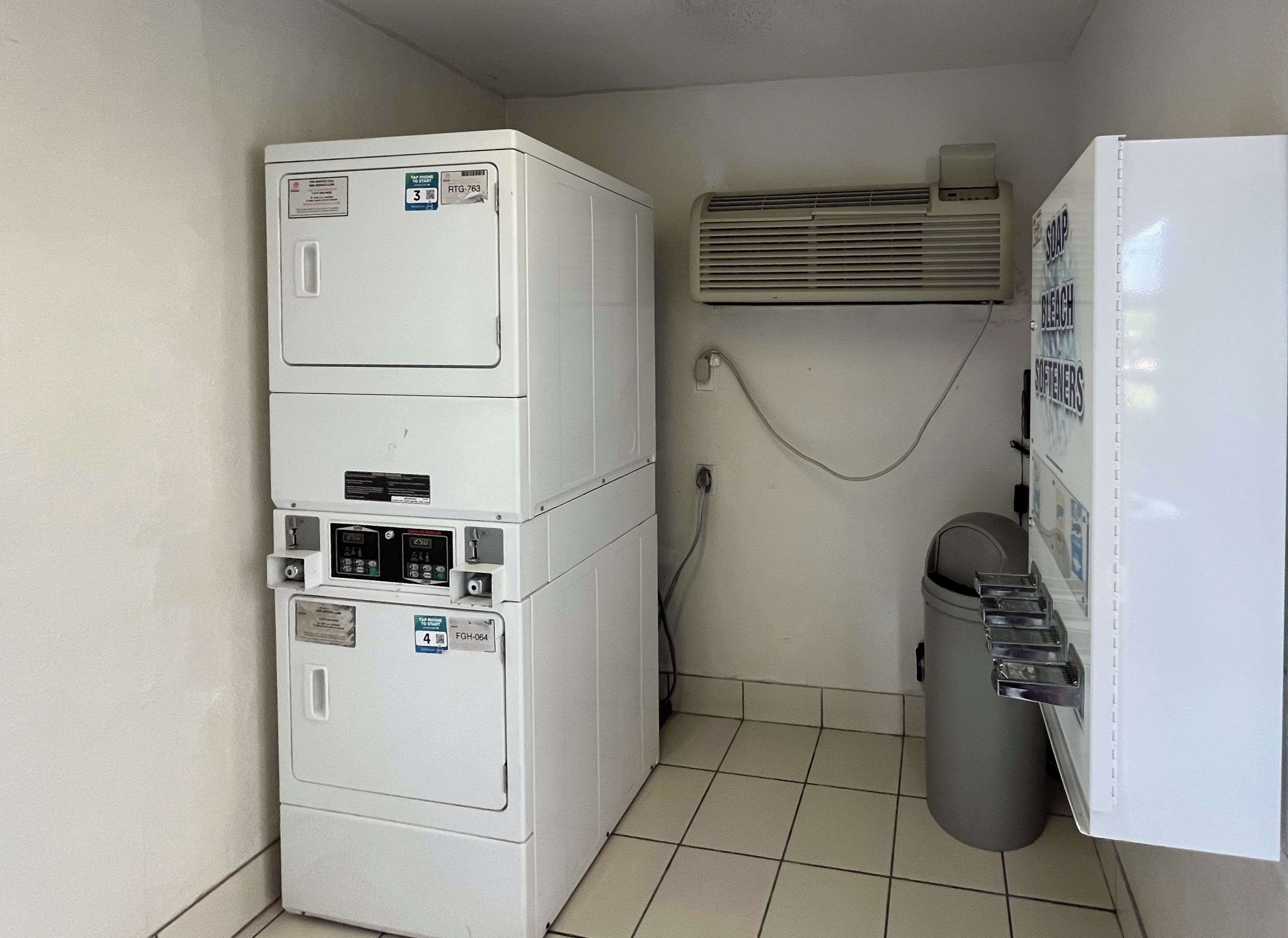 A laundry room with stacked washers and dryers, a wall-mounted air conditioner, and a trash bin.
