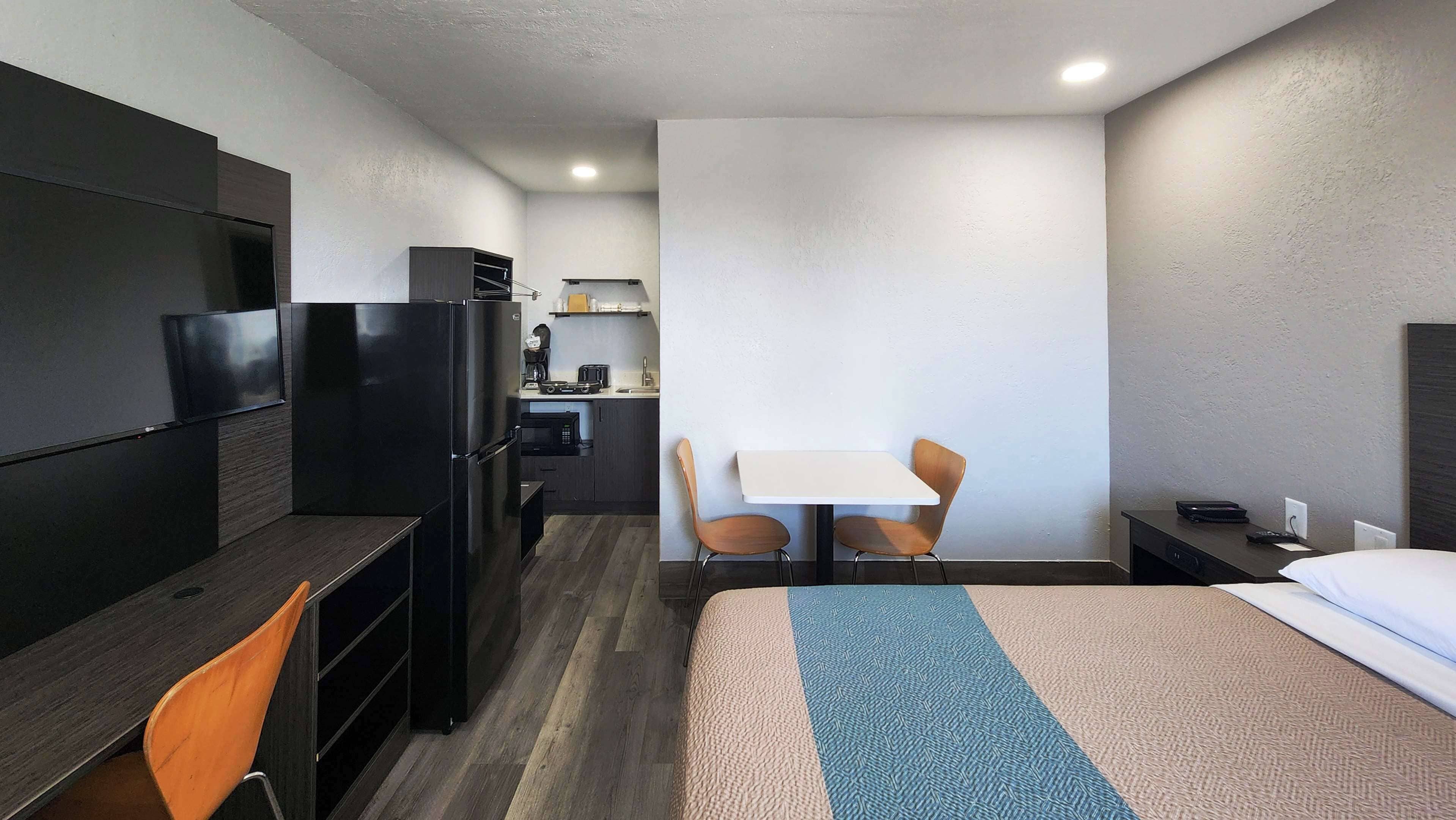 A hotel room with a bed, desk, chairs, a refrigerator, a microwave, and a small kitchenette.