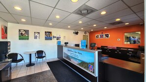 A hotel reception area with a counter, chairs, and a large screen displaying a beach scene. - Studio 6 Webster, TX Houston - NASA Lake (Webster)
