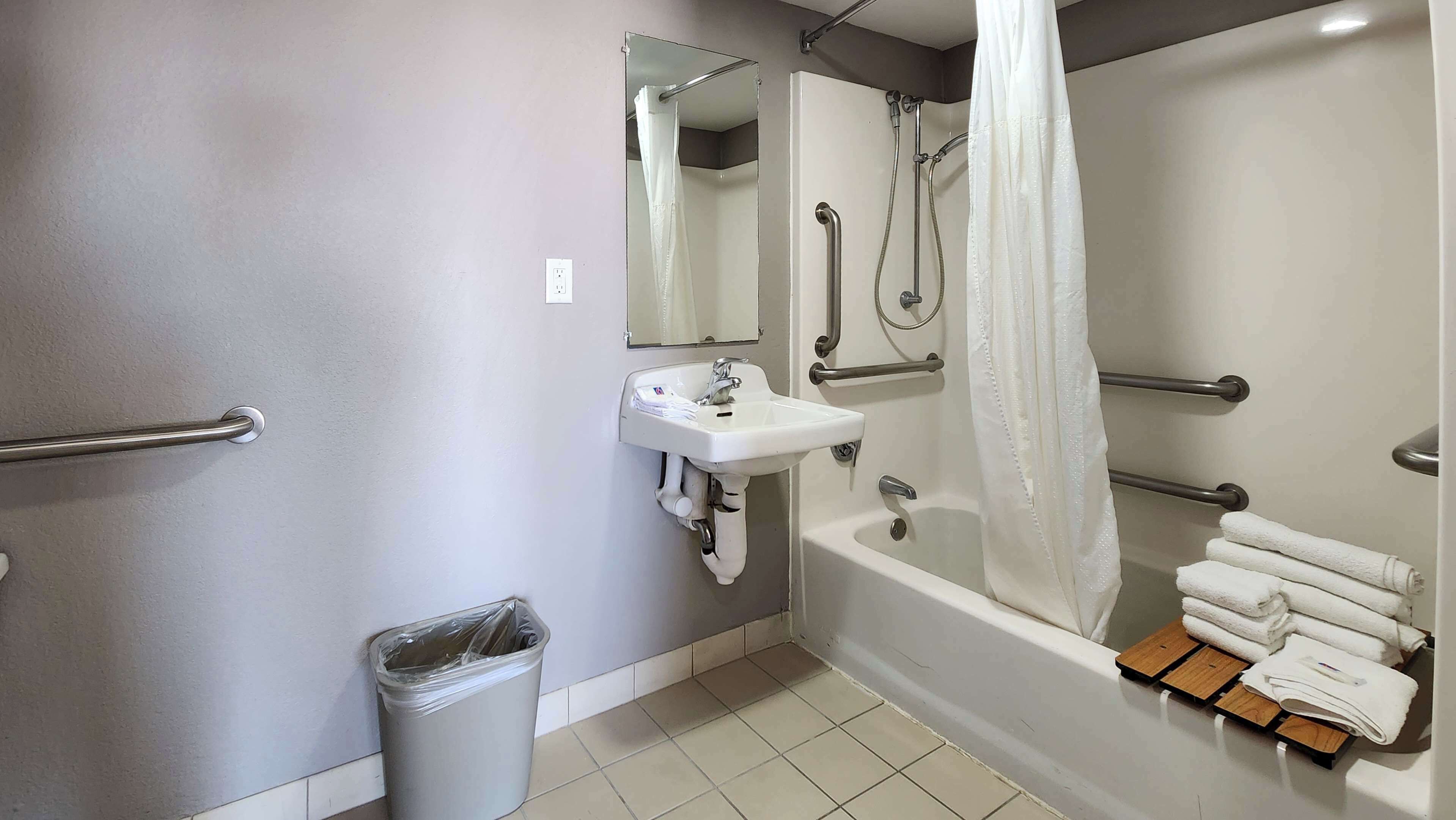 A bathroom with a sink, mirror, shower, and handrails.