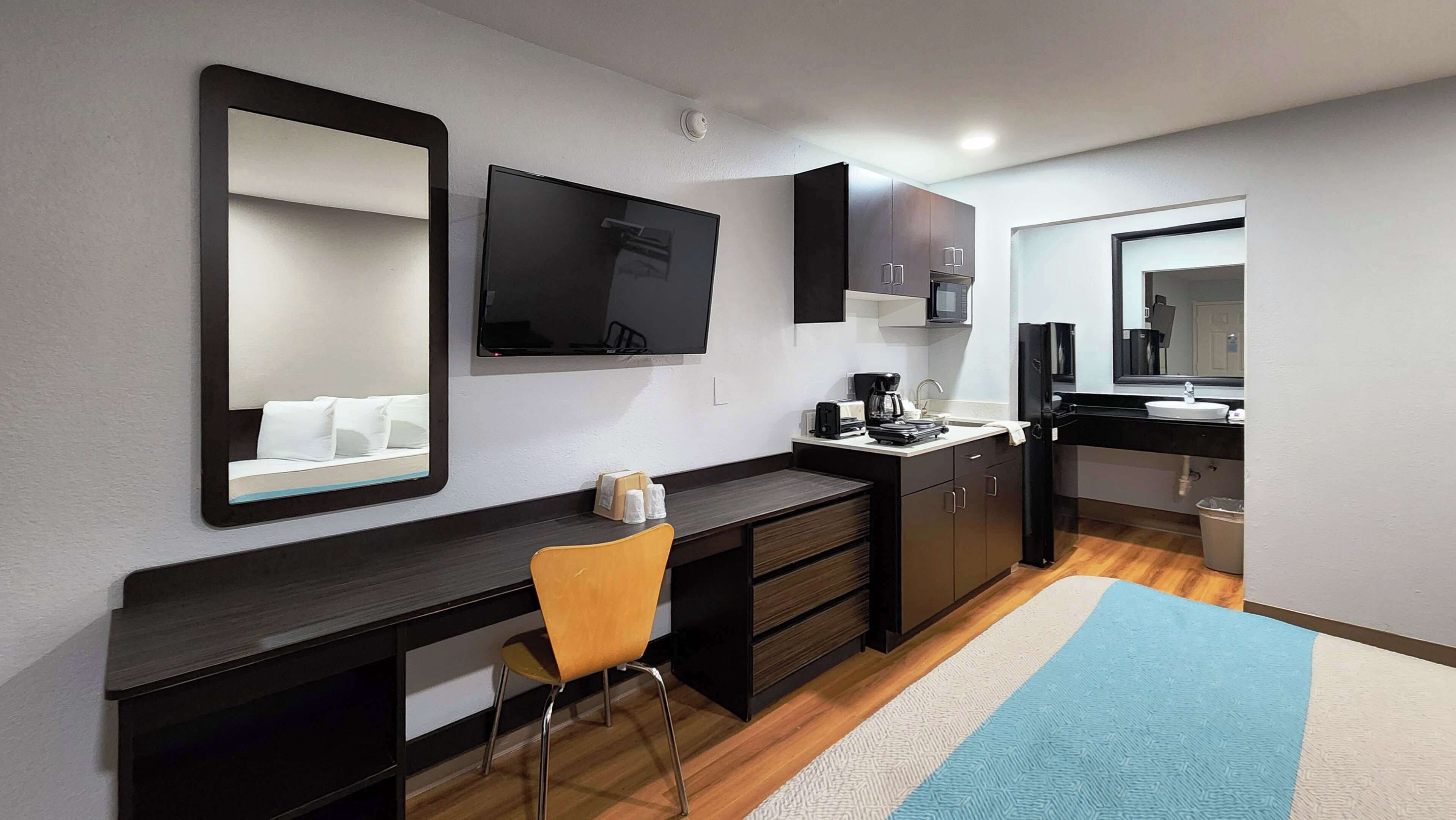 Habitación de hotel compacta con escritorio de madera oscura, una sola silla amarilla, televisor de pantalla plana montado en la pared, kitchenette pequeña con microondas y un espejo grande sobre el lavabo.