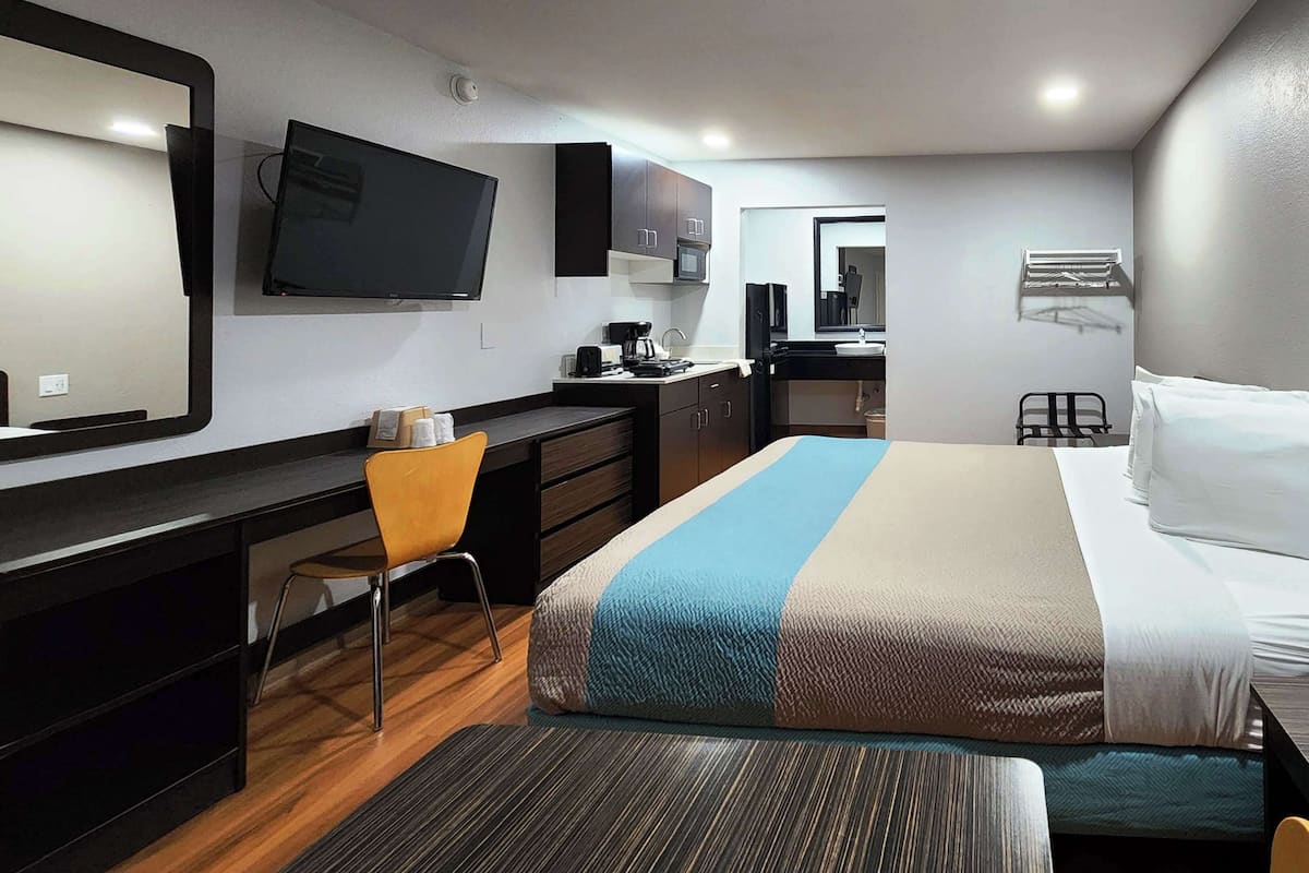 A hotel room with a bed, a desk, a chair, a TV, a microwave, a sink, and a mirror.