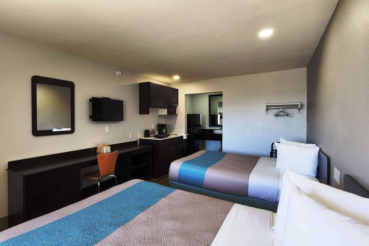 A hotel room with two beds, a desk, a chair, a television, a microwave, and a small kitchenette.
