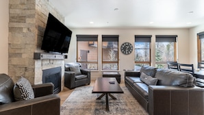 A modern living room with a stone fireplace, leather sofas, a flat-screen TV, and large windows offering a view of the outside. - Open-Concept Townhome Next to Blue River w/ Private Wrap-Around Patio! (Silverthorne)