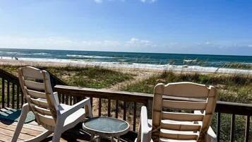A wooden deck with two rocking chairs and a small table, overlooking a beach with waves.