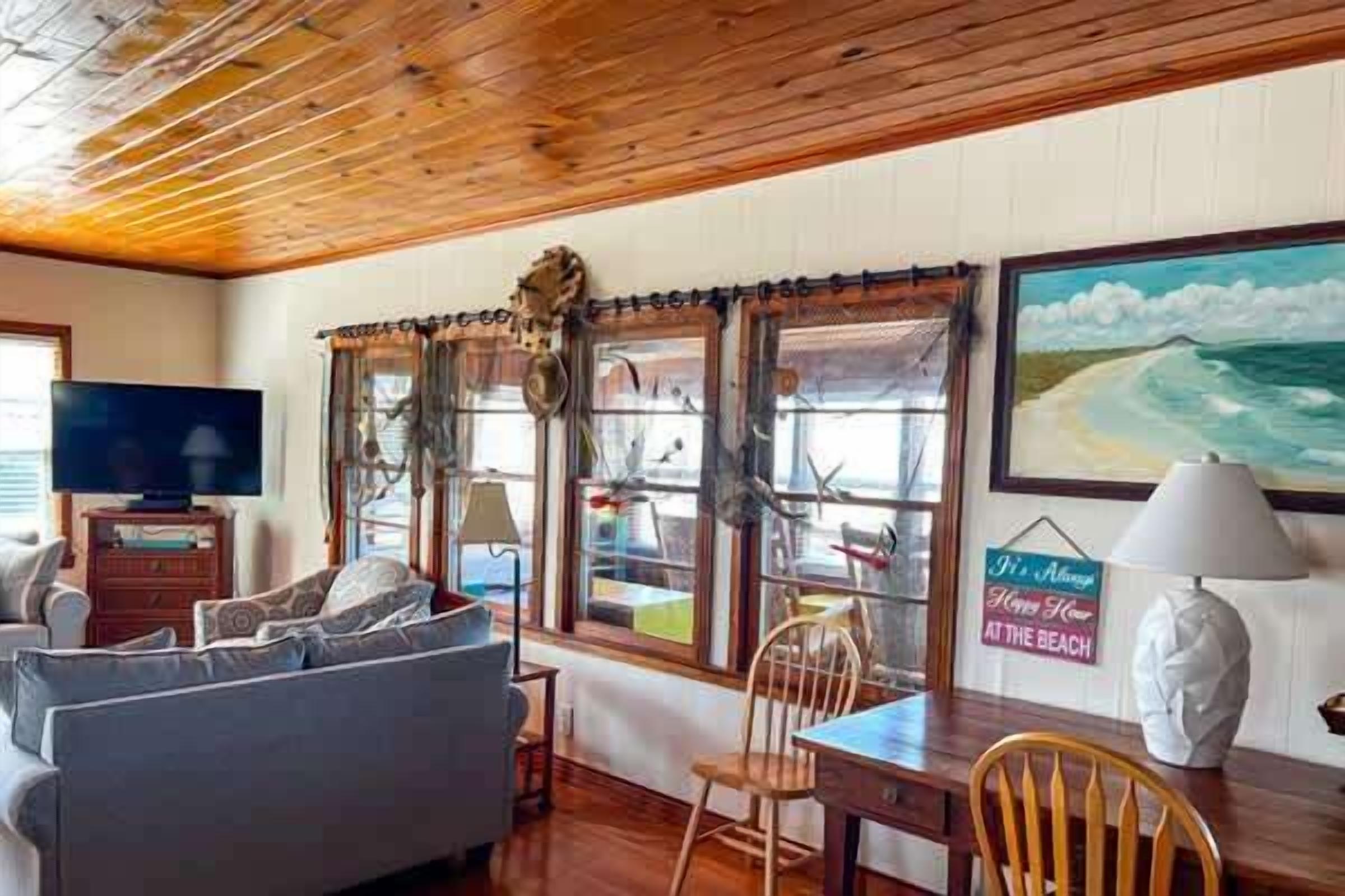 A room with a wooden ceiling, a sofa, a table, chairs, a television, and a painting of a beach scene.