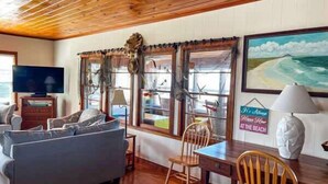 Une pièce avec un plafond en bois, un canapé, une table, des chaises, un téléviseur et un tableau représentant une scène de plage.