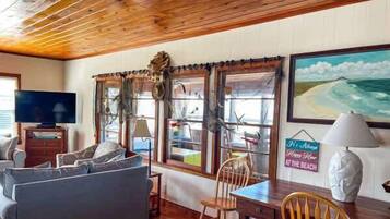 A room with a wooden ceiling, a sofa, a table, chairs, a television, and a painting of a beach scene.