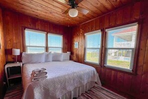 A wooden-paneled room with a bed, two windows, a ceiling fan, and a nightstand. - Beachfront Coastal Cottage St George Island (St.George Island)