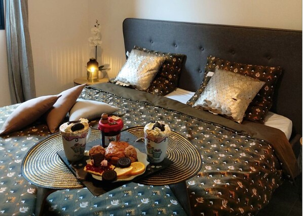 Un lit avec une tête de lit sombre et plusieurs oreillers, une table de chevet sur laquelle se trouve un plateau avec des pâtisseries et des boissons, et une lampe sur la table de chevet.