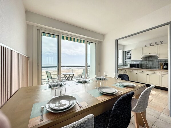 A dining area with a view of the beach and ocean, a kitchen with white cabinets, and a dining table set with plates and glasses.