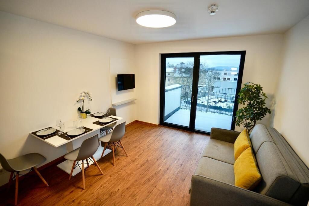 Una sala de estar moderna con comedor, un sofá con cojines amarillos, un ventanal con vistas a edificios y un televisor de pantalla plana montado en la pared.