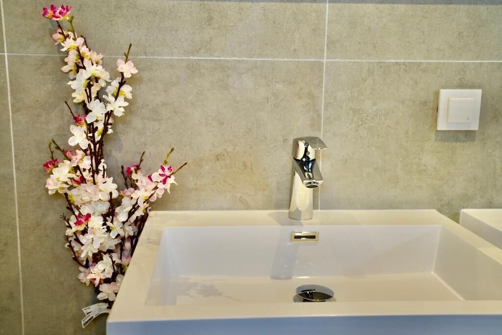 A bathroom with a white sink, a chrome faucet, and a decorative branch of flowers.