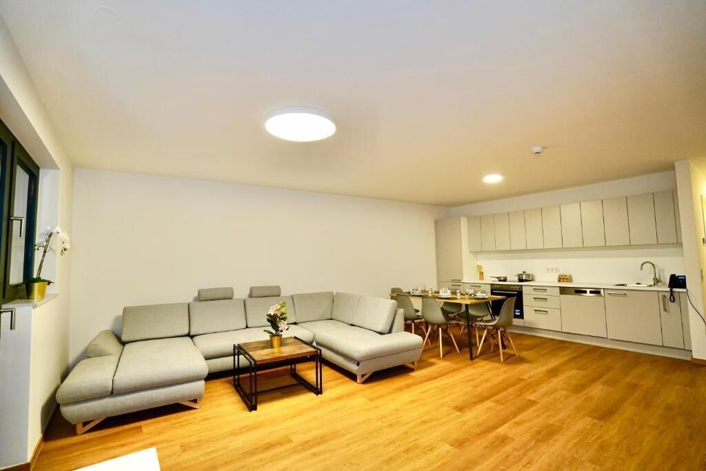A modern living room with a sectional sofa, a coffee table, and a dining area with a small table and chairs. The kitchen is visible with white cabinets and a countertop.