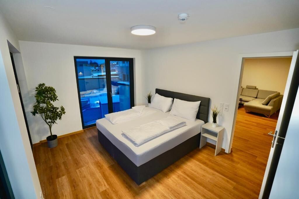 Ein modernes Schlafzimmer mit einem großen Bett, einem kleinen Nachttisch, einer Zimmerpflanze und einer Glastür, die auf einen Balkon führt.