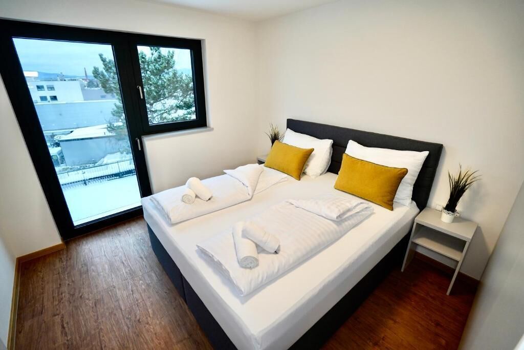 Een moderne slaapkamer met een groot bed, een nachtkastje, een raam met uitzicht op gebouwen en een kleine plant op het nachtkastje.