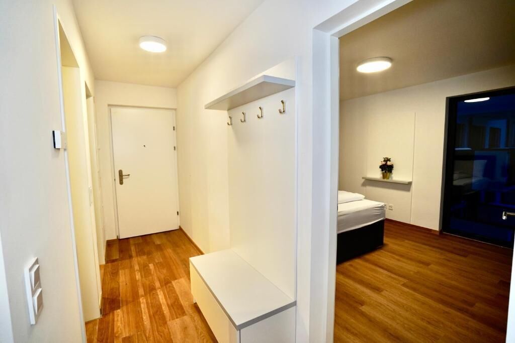 Une pièce moderne et bien éclairée, dotée d’un plancher en bois, d’un banc blanc, d’un lit et d’une porte donnant sur une autre pièce.
