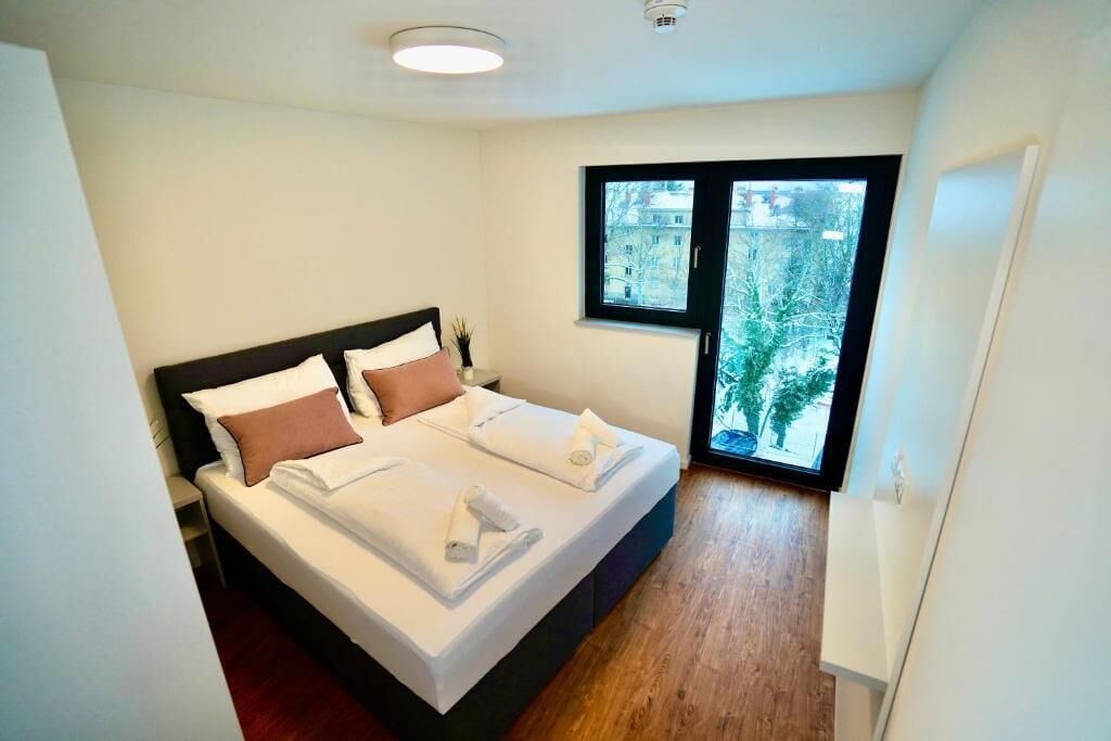 A modern bedroom with a large bed, a window with a view of trees, and a small bedside table.