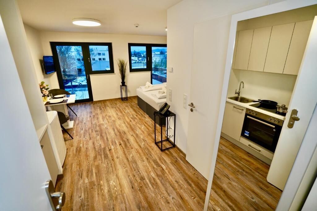 A compact apartment with a wooden floor, a kitchenette, a small seating area, and large windows offering a view of the outside.