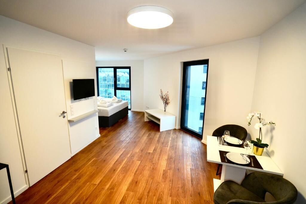 Een moderne woonkamer met een houten vloer, een witte bank, een aan de muur bevestigde televisie en een eethoek met een tafel gedekt voor twee personen.