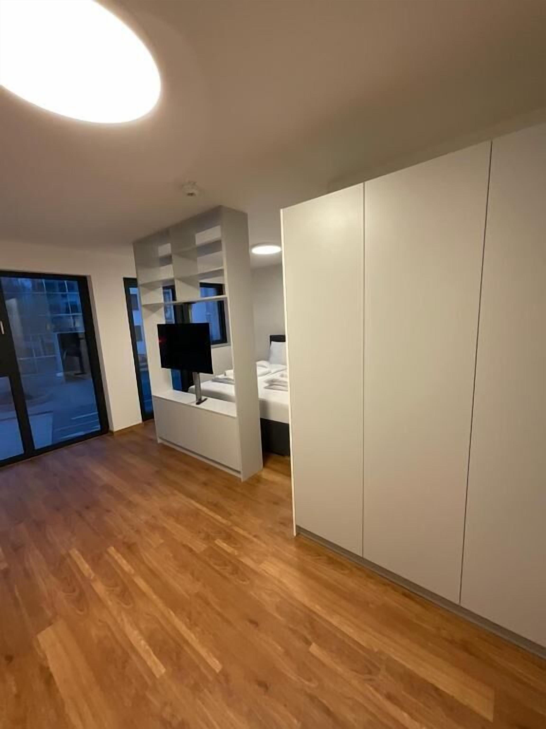 A modern hotel room with a wooden floor, a large wardrobe, a bed, a TV, and a window with a view of the outside.