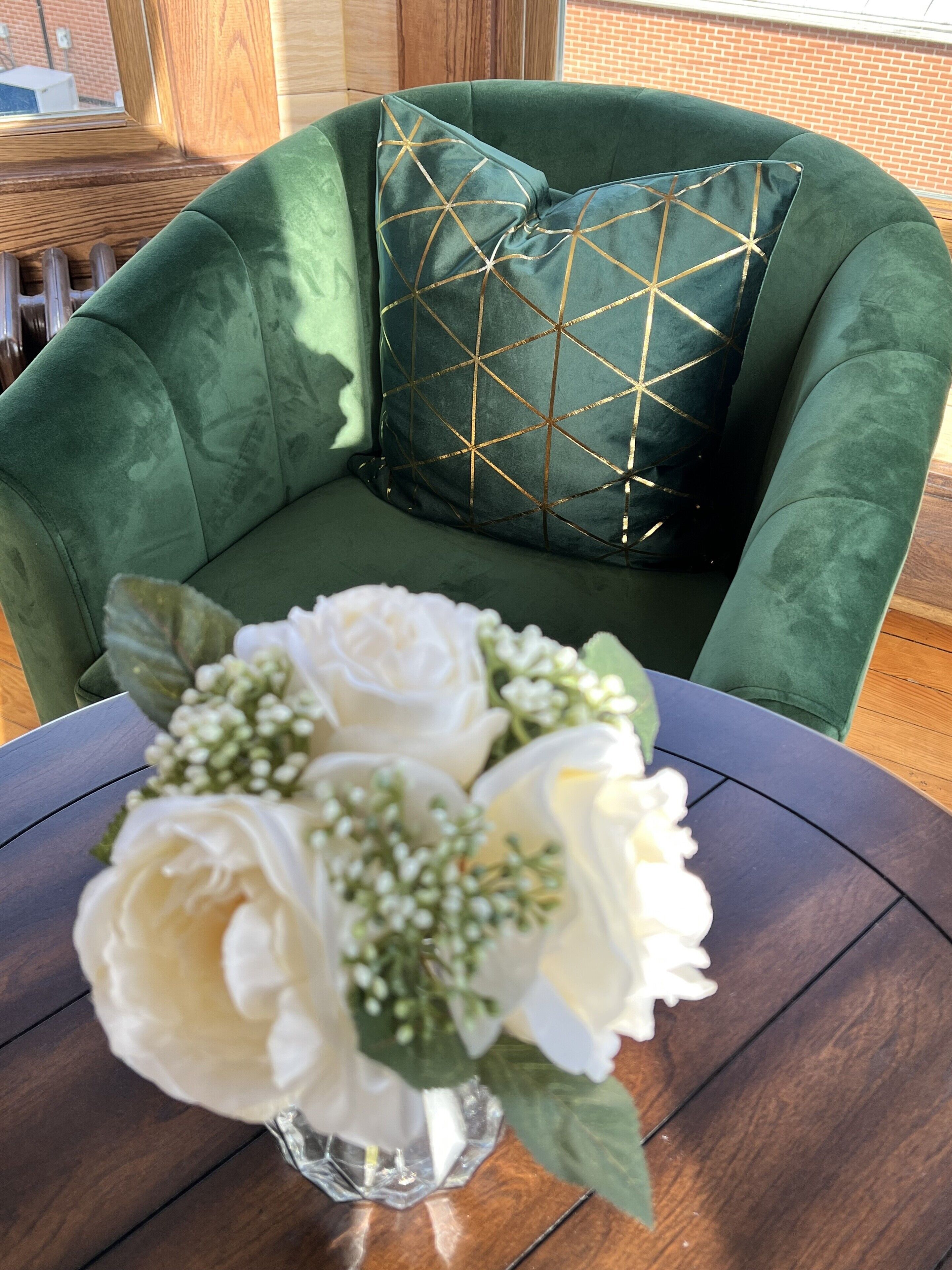 A green upholstered chair with a geometric patterned pillow, a round table with a vase of white flowers, and a brick wall in the background.