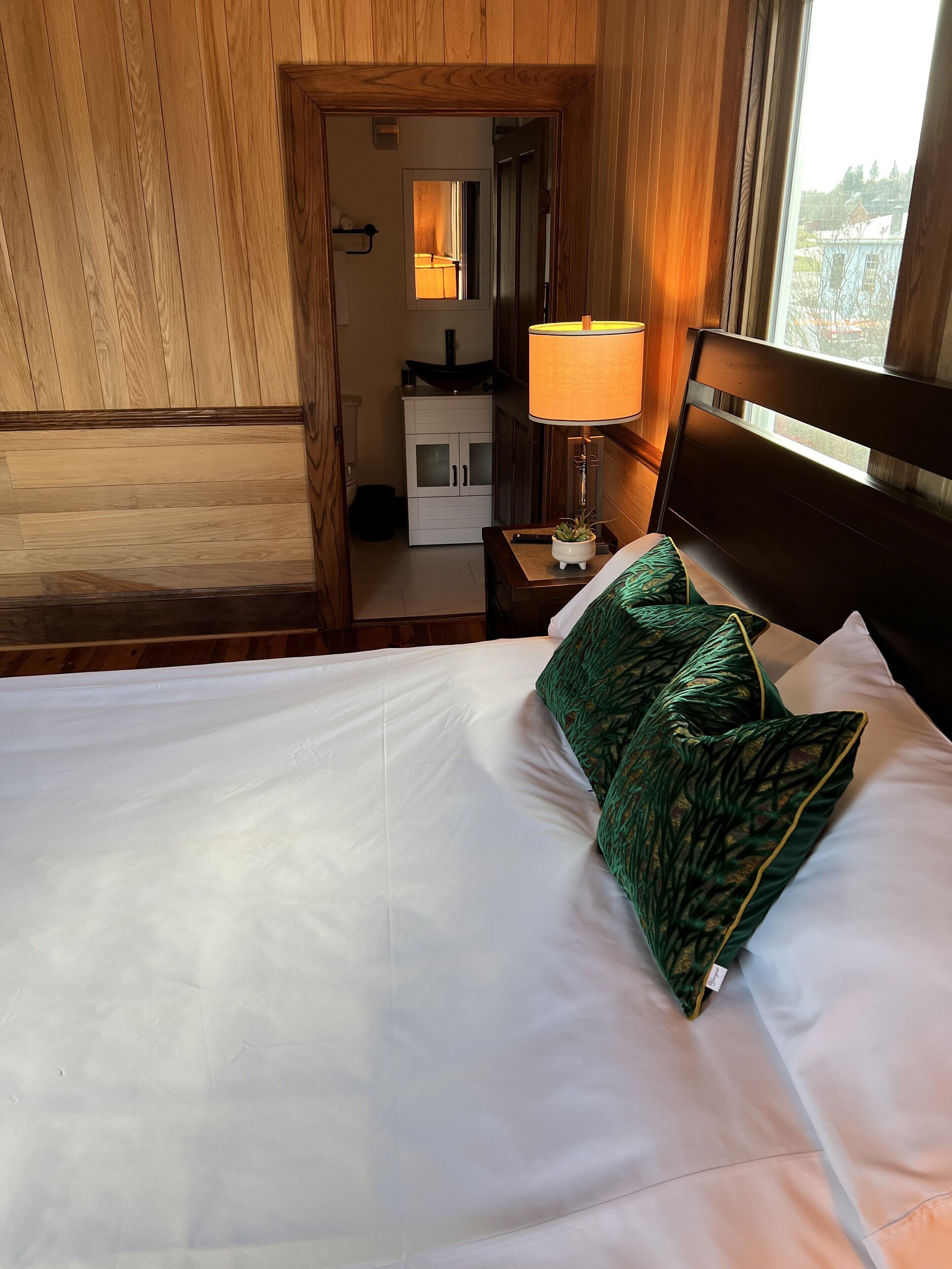 Habitación de hotel con pared revestida de madera, cama con ropa blanca y almohadas de diseño verde, mesita de noche con lámpara y vista al exterior por una ventana.