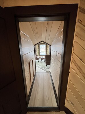 A narrow wooden hallway with a sloped ceiling leading to a room with a window and two chairs.