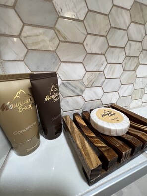 A bathroom vanity with a hexagonal tile backsplash, a bottle of Mountain Escape conditioner, a tube of Mountain Escape shampoo, and a bar of Mountain Escape luxury soap.