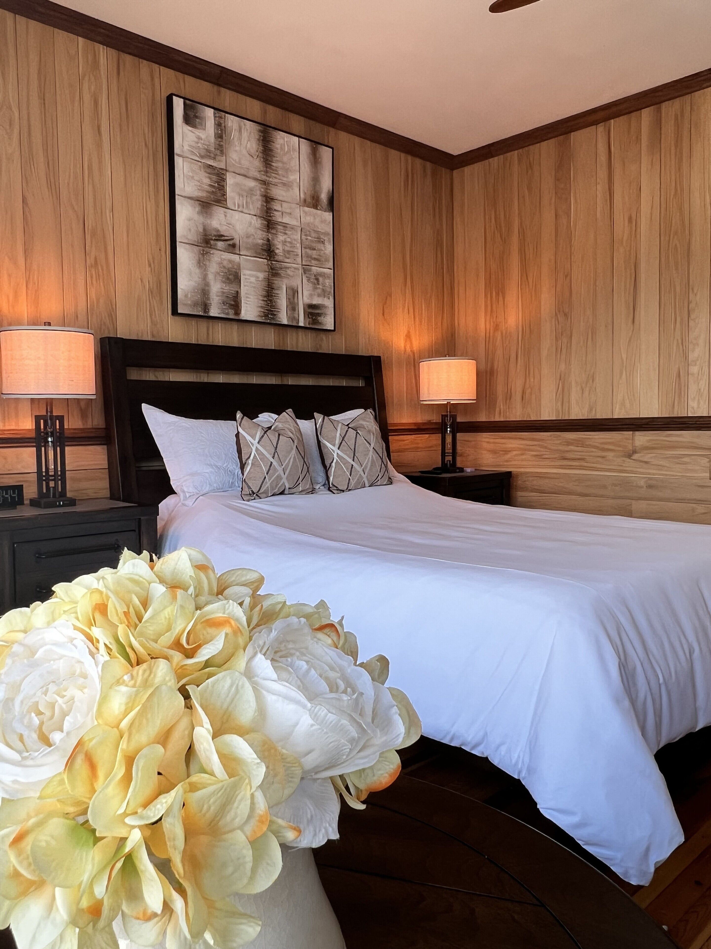 A hotel room with a large bed, wooden panel walls, two bedside lamps, and a large floral arrangement.