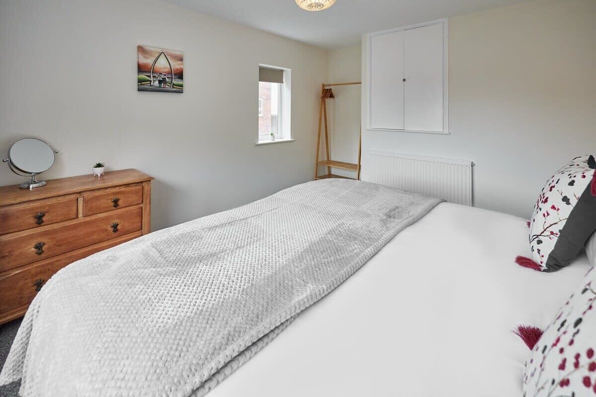 A bedroom with a bed, a wooden dresser, a bedside table with a mirror, a window, a radiator, and a wall-mounted wardrobe.