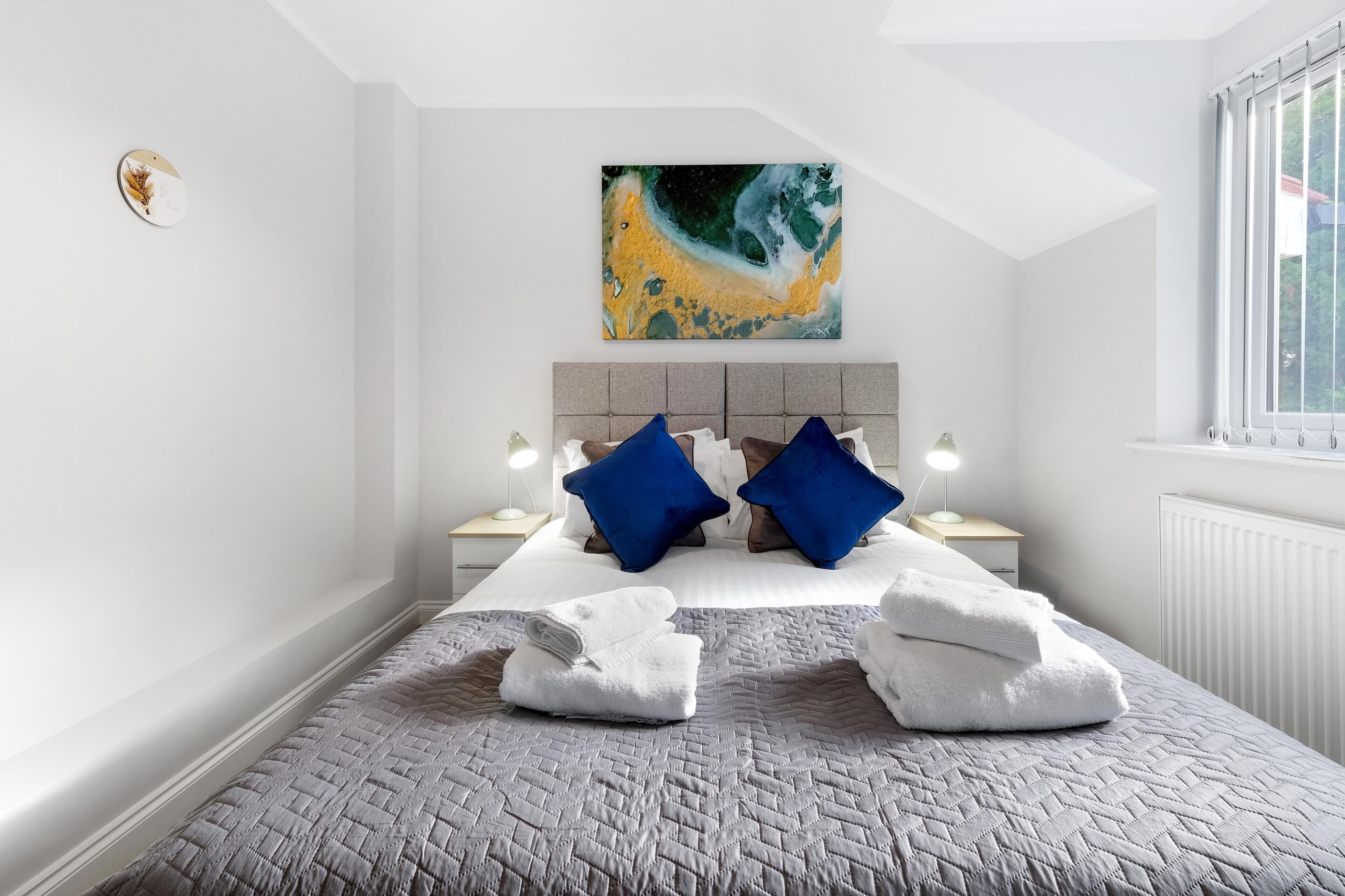 A modern bedroom with a large bed, two blue pillows, a gray quilted bedspread, and a painting above the bed.