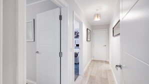 Un couloir aux murs blancs, doté d’un plancher en bois et d’une porte donnant sur une chambre à coucher, où se trouvent un lit et un tapis bleu.