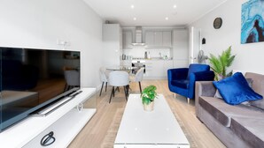 Un salon moderne avec un téléviseur à écran plat, une table basse agrémentée d’une plante, un canapé gris avec des coussins bleus et un coin repas avec une table et des chaises en verre.