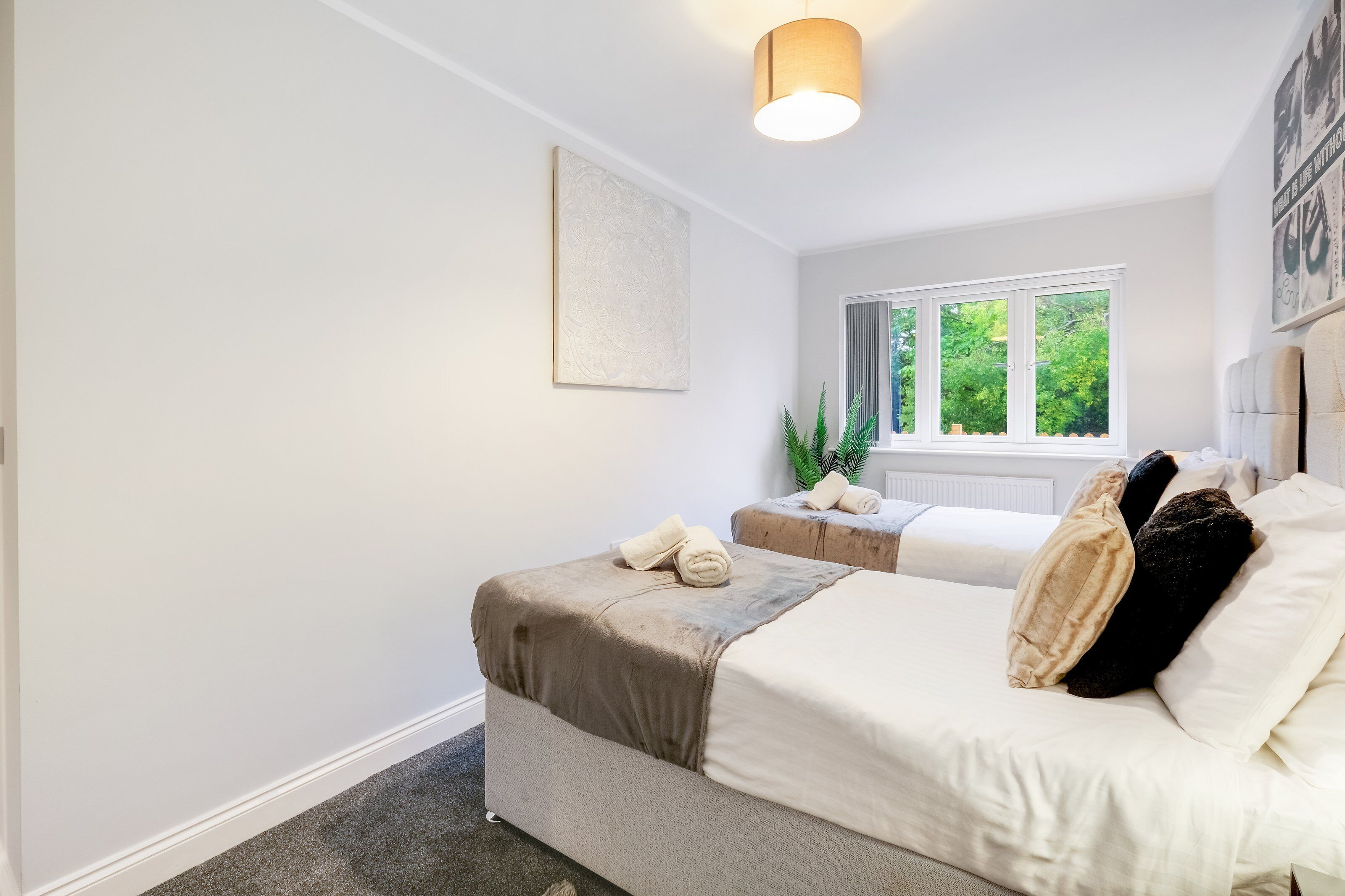 Une chambre avec deux lits, une fenêtre donnant sur un paysage verdoyant, un luminaire au plafond et une œuvre d’art accrochée au mur.