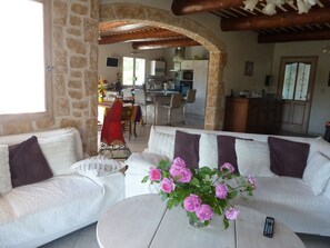 Ein gemütliches Wohnzimmer mit einem weißen Sofa, einem runden Couchtisch und einer Vase mit rosa Blumen. Ein Bogen führt in eine Küche und einen Essbereich.
