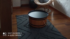 Una habitación con piso de madera, una silla de mimbre, una mesa redonda, una ventana con cortinas y un cuadro enmarcado en la pared.
