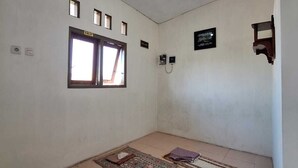 Une pièce avec un sol carrelé, une fenêtre avec des rideaux, un luminaire fixé au mur et une petite étagère sur laquelle est posé un objet encadré.