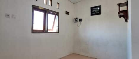 A room with a tiled floor, a window with curtains, a wall-mounted light fixture, and a small shelf with a framed item.