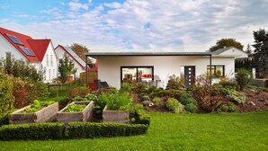 Une maison moderne avec un toit rouge, des panneaux solaires et un jardin bien entretenu, agrémenté de plates-bandes et de diverses plantes.