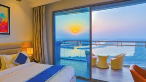 Habitación de hotel con un balcón grande, una cama con almohadas, una mesita de noche con lámpara y vistas al atardecer sobre el agua.