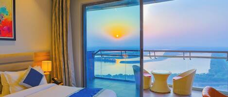 Ein Hotelzimmer mit einem großen Balkon, einem Bett mit Kissen, einem Nachttisch mit Lampe und Blick auf den Sonnenuntergang über dem Wasser.