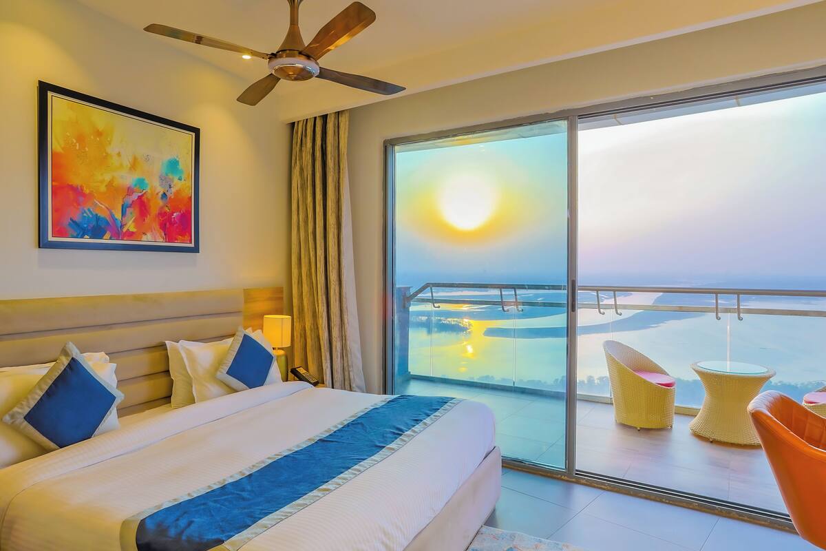 A hotel room with a large bed, a ceiling fan, a painting, a view of the sea, and a balcony with chairs.