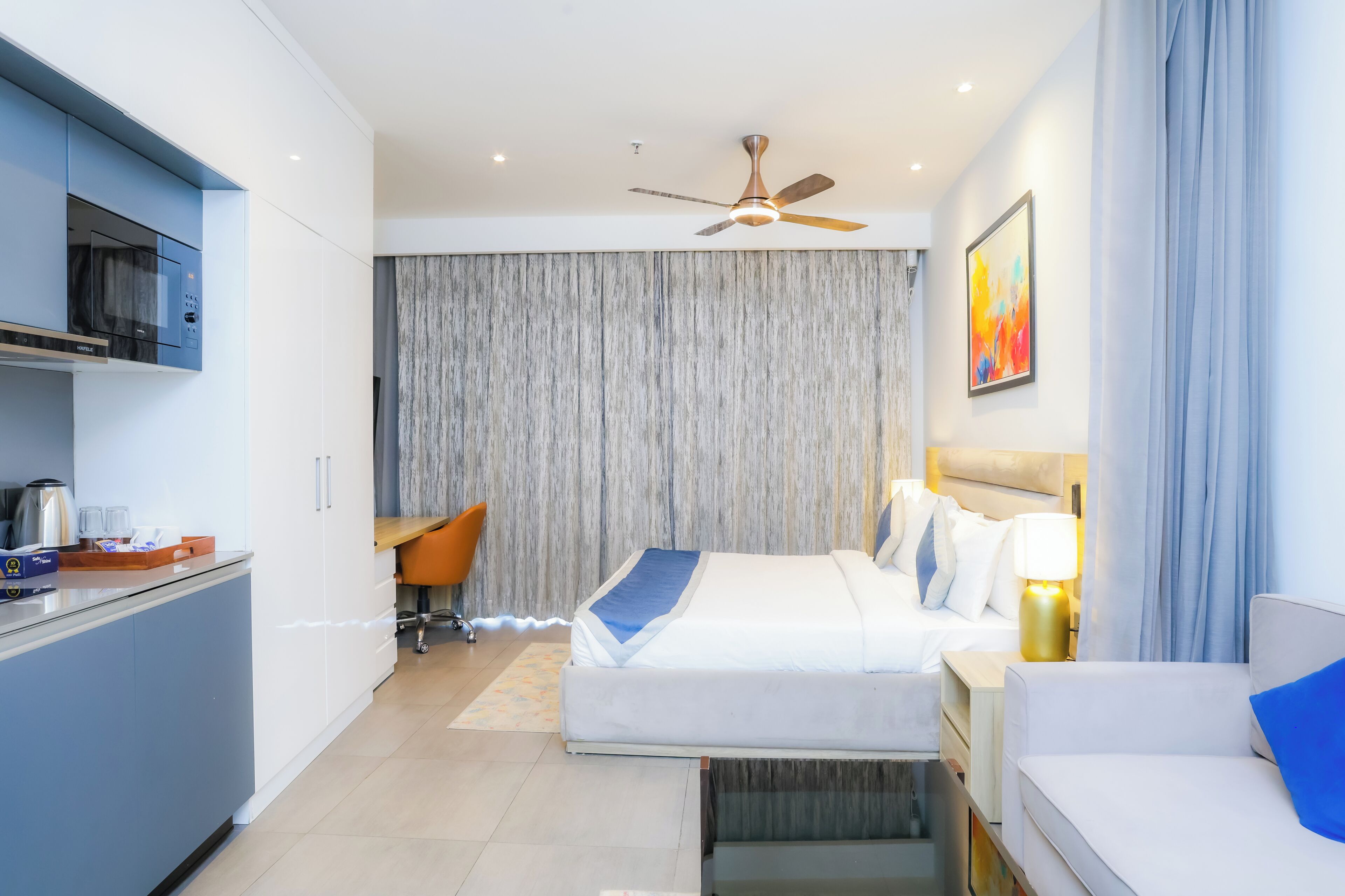 A modern hotel room with a bed, a desk, a kitchenette, and a ceiling fan.