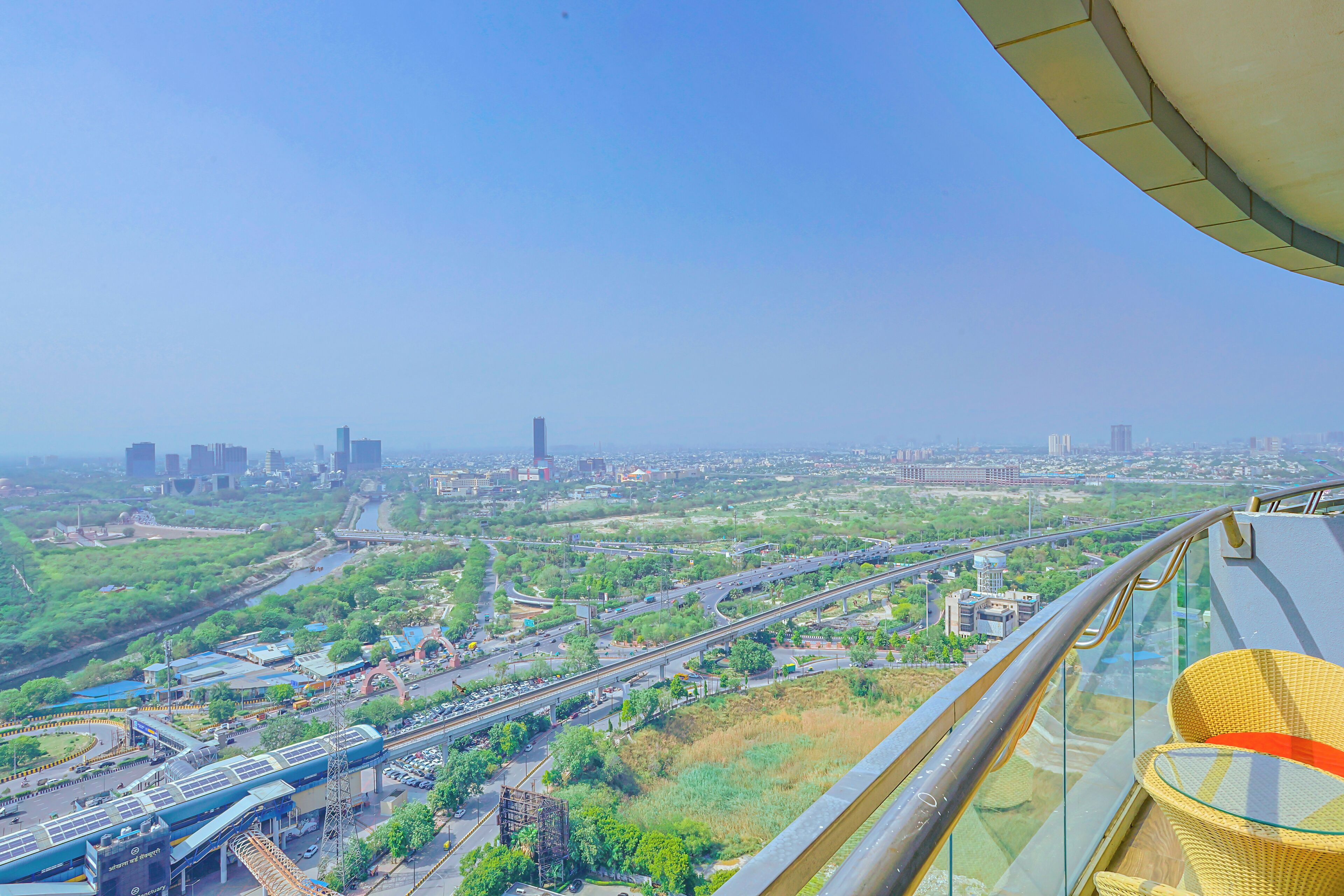 A high-rise building with a glass balcony overlooking a cityscape, including roads, greenery, and buildings.