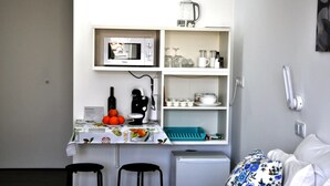 A compact kitchenette with a microwave, coffee maker, and a small refrigerator. There are two black chairs, a table with a fruit bowl, and a bed with floral bedding. - Cozy Seaside Studio Apartment C - Faraway (Lagoa)