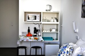 Eine kompakte Küchenzeile mit einer Mikrowelle, einer Kaffeemaschine und einem kleinen Kühlschrank. Zwei schwarze Stühle, ein Tisch mit Obstschale und ein Bett mit floralem Bettbezug.